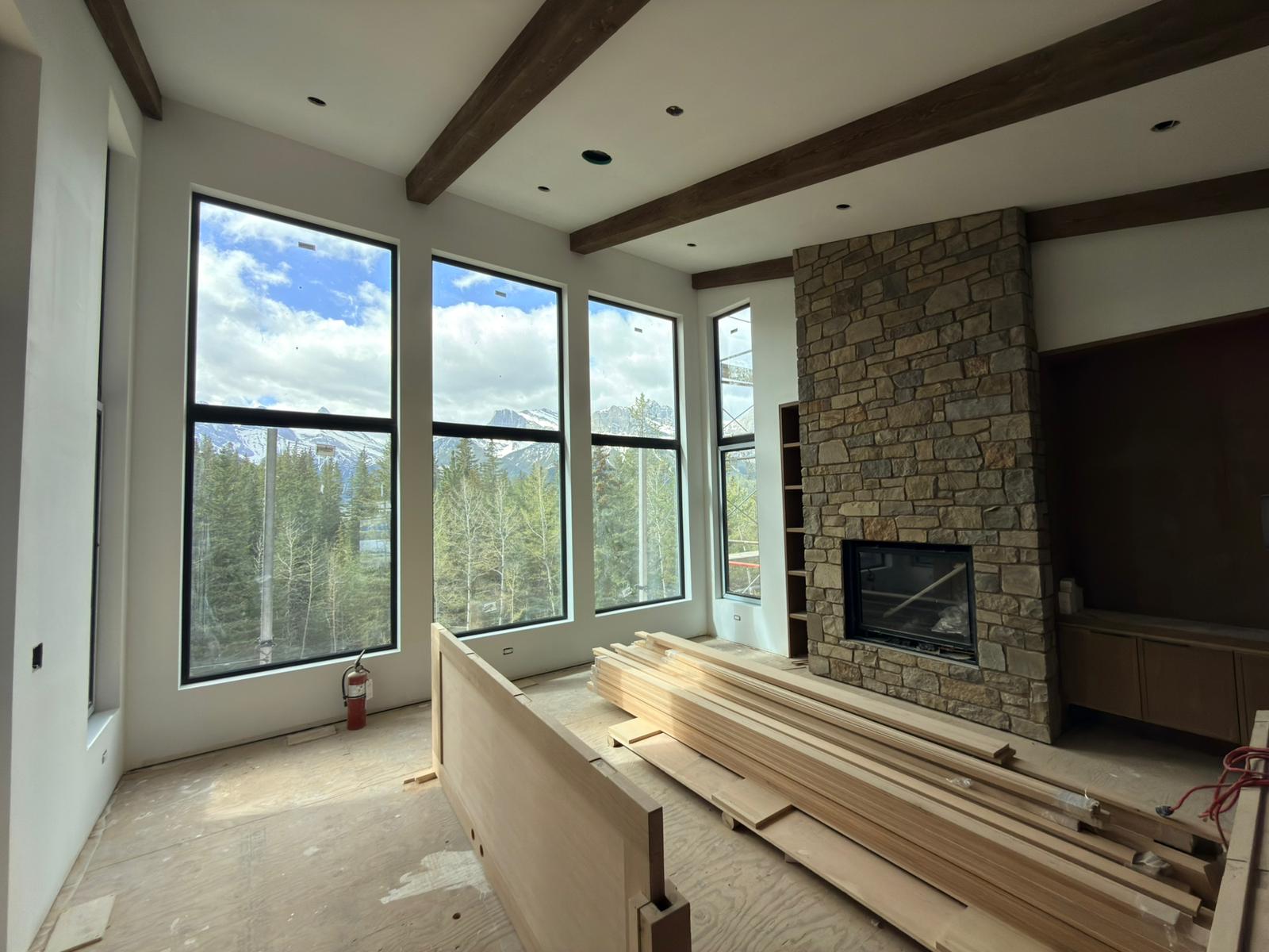 Acreage great room near Calgary — five tall black-frame REHAU windows opening onto a Rocky Mountain view, stone fireplace, exposed beams